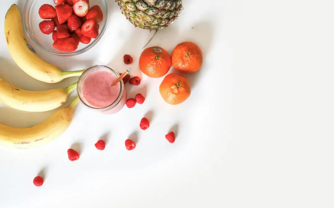 Jasne, świeże owoce – banany, truskawki, ananasy i pomarańcze – otaczają szklankę różowego smoothie na białym tle, przywodząc na myśl radosną, zdrową przekąskę.